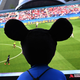 Uma pessoa usando orelhas de mickey mouse assiste a uma tela gigante no meio de uma rua mostrando um jogo de futebol.